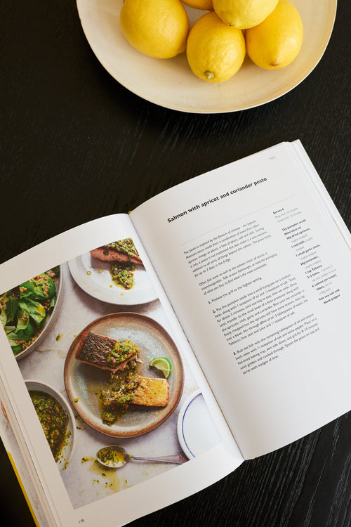 An open cookbook spread on a dark wooden table, showing a recipe page for salmon with asparagus and citrus pistachio. A plate of lemons sits in the background.