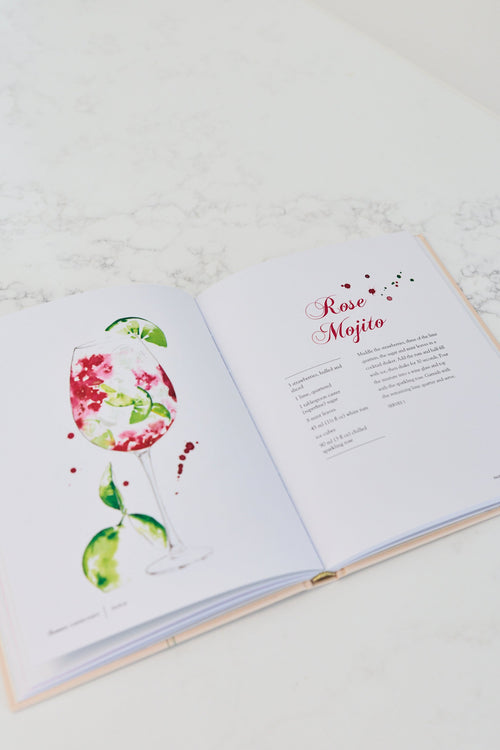 An open hardcover recipe book on a light marble surface, showing a watercolor illustration of a rose mojito on the left page and the recipe title 'Rose Mojito' on the right page.