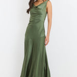 A woman modeling a sleeveless olive green satin maxi dress with a cowl neckline and a flared skirt, standing against a plain white studio background.
