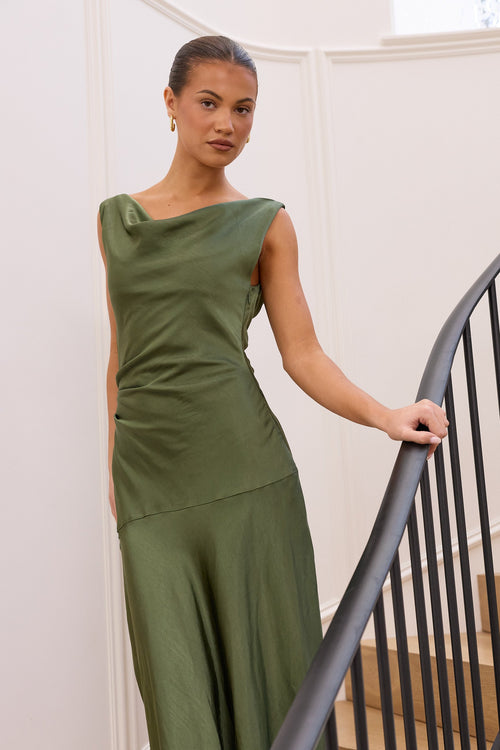 A woman wearing a sleeveless olive green maxi dress stands on an indoor staircase, holding the black metal railing. The dress has a draped, asymmetrical neckline and a flowing silhouette.