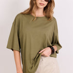 A woman wearing an oversized olive green short-sleeve t-shirt and a beige mini skirt, styled with dark sunglasses and bracelets, against a plain light gray background.