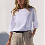 A woman wearing a white off-the-shoulder top, a beige linen mini skirt, a black belt, and black sandals, standing in a sunny, minimalist white-walled courtyard.