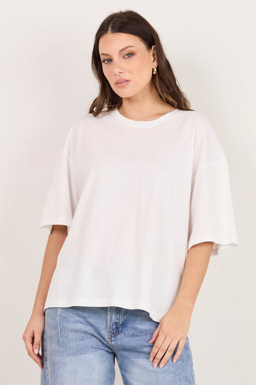 A woman modeling a loose white short-sleeve t-shirt and light-wash blue cropped jeans with black heeled sandals, standing against a plain off-white studio background.