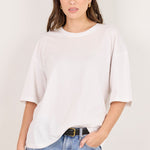 A woman modeling an oversized white short-sleeve crewneck t-shirt paired with light-wash blue jeans and a black belt, standing against a plain off-white studio background.