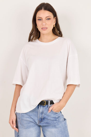 A woman modeling an oversized white short-sleeve crewneck t-shirt paired with light-wash blue jeans and a black belt, standing against a plain off-white studio background.