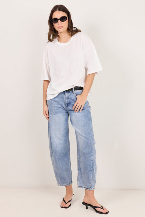 A woman modeling a white oversized short-sleeve t-shirt, light-wash blue cropped jeans, a black belt, and black heeled sandals against a plain off-white studio background.