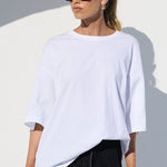 A woman modeling a white oversized short-sleeve top and black wide-leg lace pants, standing in a bright white architectural setting with a curved wall and shadow. She is wearing black sunglasses and holding a red handbag.