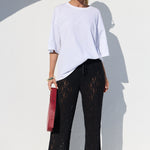 A model stands in a white architectural setting wearing a white oversized t-shirt, black lace wide-leg pants, and holding a red clutch bag.