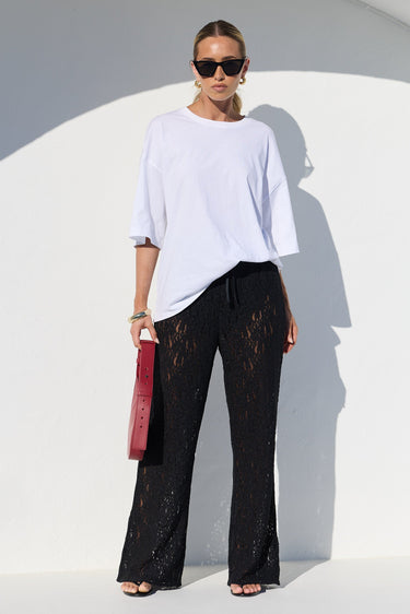 A model stands in a white architectural setting wearing a white oversized t-shirt, black lace wide-leg pants, and holding a red clutch bag.