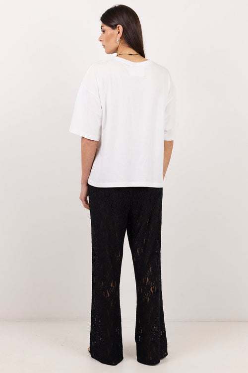 A woman is shown from the back wearing a white short-sleeve crop top and black wide-leg pants with a textured lace-like pattern, standing against a plain white studio background.