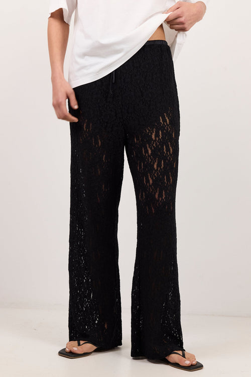 A person wearing loose black crochet or lace wide-leg pants with a white top and black sandals, standing against a plain light studio background.