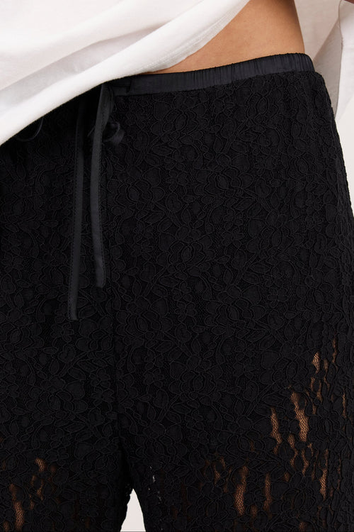 Close-up of a person wearing black lace pants with a sheer openwork pattern and a black drawstring waist, while lifting a white shirt to show the fit.