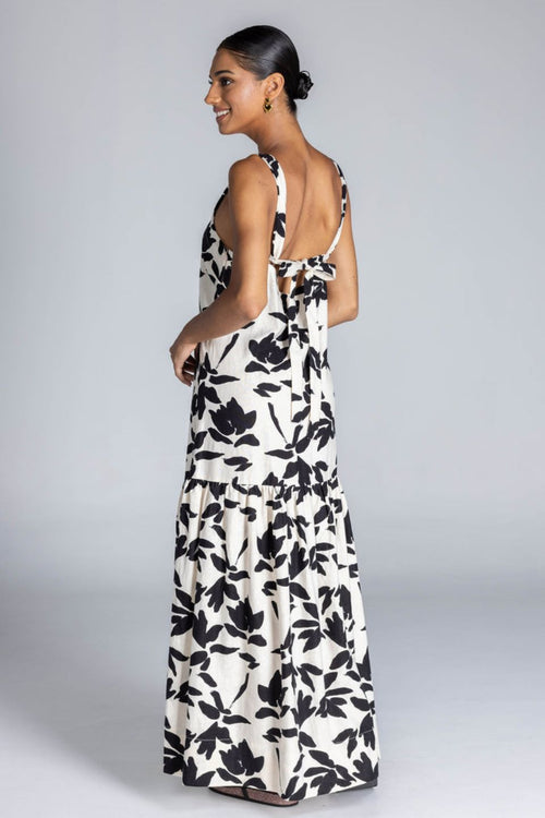 Woman wears an ivory maxi dress with a bold black floral print, wide straps, V-neckline, back tie detail, and a flowing skirt with side splits. The look is fresh, feminine, and relaxed.
