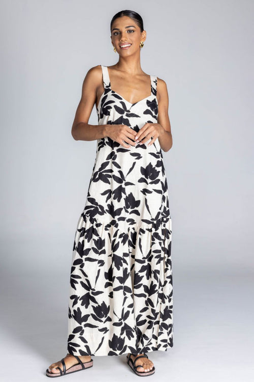 Woman wears an ivory maxi dress with a bold black floral print, wide straps, V-neckline, back tie detail, and a flowing skirt with side splits. The look is fresh, feminine, and relaxed.
