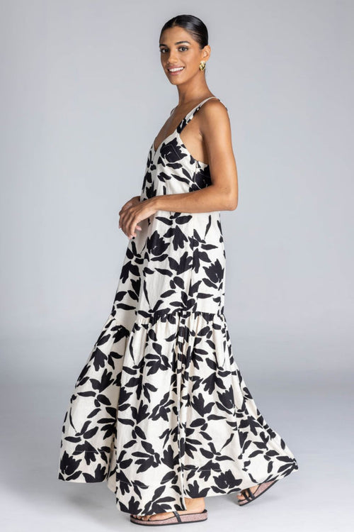 Woman wears an ivory maxi dress with a bold black floral print, wide straps, V-neckline, back tie detail, and a flowing skirt with side splits. The look is fresh, feminine, and relaxed.