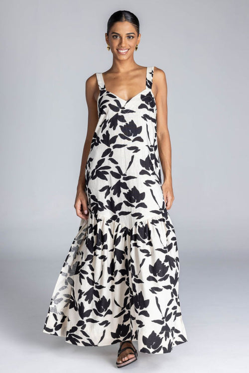 Woman wears an ivory maxi dress with a bold black floral print, wide straps, V-neckline, back tie detail, and a flowing skirt with side splits. The look is fresh, feminine, and relaxed.