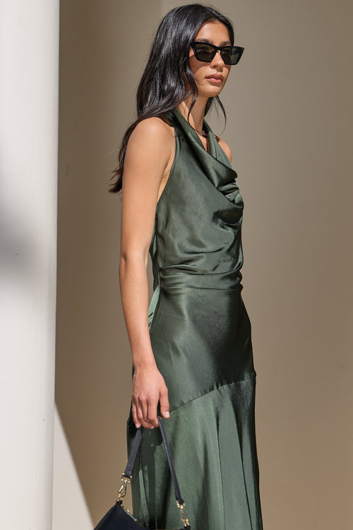 A woman wearing a dark olive green satin halter dress and black sunglasses, holding a navy blue handbag, standing in front of a beige wall.