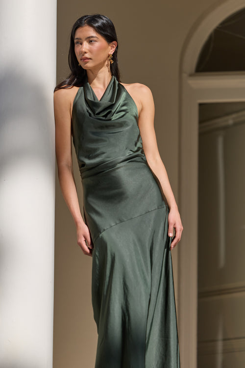A woman wearing a dark green satin halter-neck maxi dress, standing in a softly lit interior with beige walls and an arched doorway in the background.