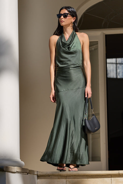 A woman wearing a dark green satin halter-neck maxi dress stands on stone steps outside a building with white columns, holding a dark blue-gray handbag.