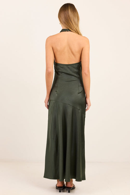 A woman viewed from the back wearing a dark olive green halter-neck maxi dress with a satin-like sheen, standing against a plain off-white studio background.