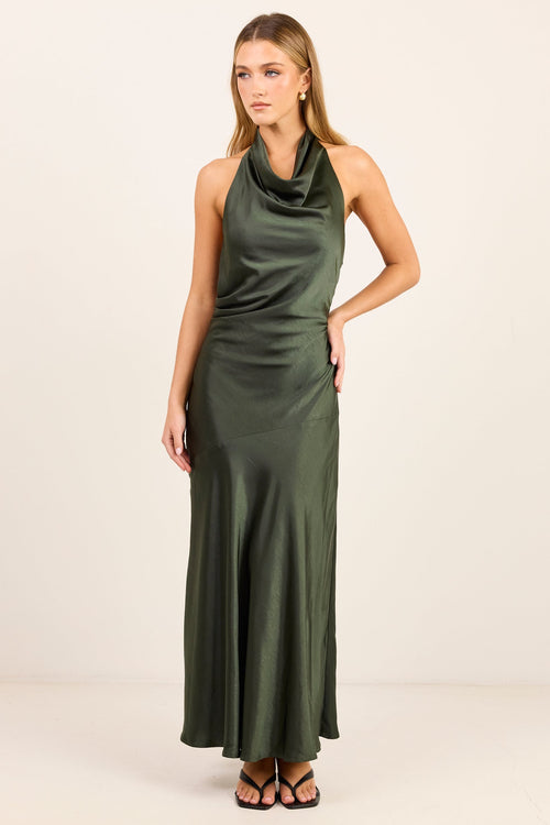 A woman modeling a sleeveless, dark olive green satin maxi dress with a draped cowl neckline, standing against a plain off-white studio background.