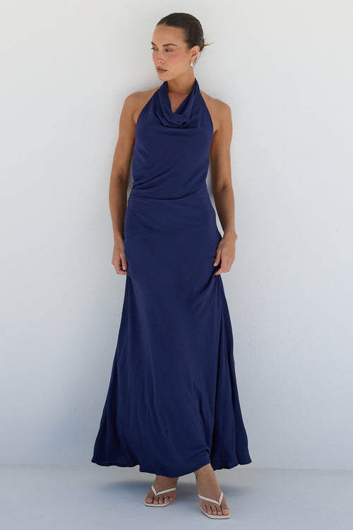 A woman modeling a sleeveless navy blue halter-neck maxi dress with a draped cowl neckline, standing against a minimalist white arched studio backdrop.
