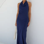 A woman wearing a sleeveless navy blue cowl-neck maxi dress stands inside a white arched alcove on a sunny day, holding a white clutch.