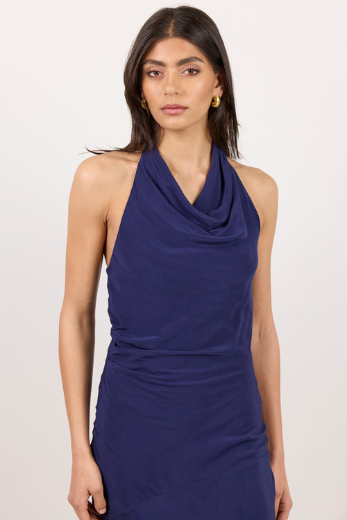 A woman modeling a sleeveless halter-neck dress in a deep navy blue color, featuring a draped cowl neckline and ruched detailing at the waist, set against a plain light background.