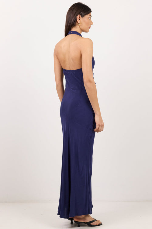 A woman modeling a navy blue halter-neck maxi dress from the back, standing against a plain white studio background.