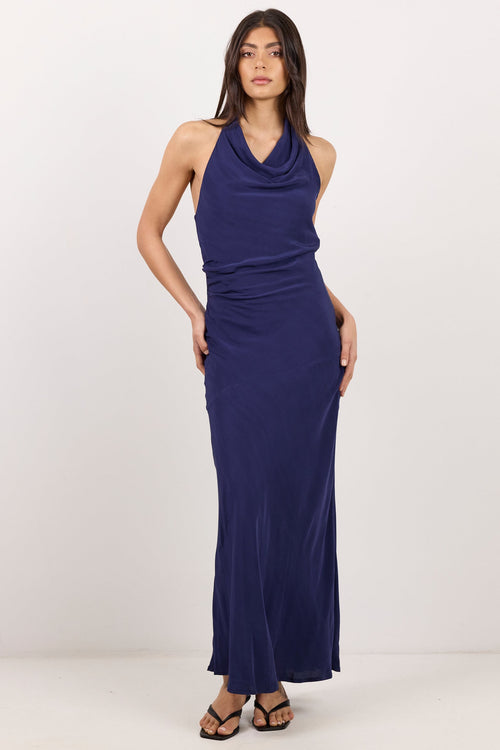 A woman modeling a sleeveless navy blue cowl-neck maxi dress with side pockets, paired with black strappy sandals, against a plain white studio background.