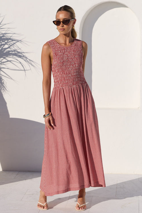 A woman modeling a sleeveless midi dress with a textured, patterned dusty rose bodice and a matching vertically striped skirt, set against a bright white stucco wall with an archway and a large terracotta planter.