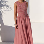 A woman modeling a sleeveless midi dress with a textured, smocked bodice and a fine vertical stripe pattern on the skirt, set against a bright white Mediterranean-style wall with an archway and a large terracotta pot.