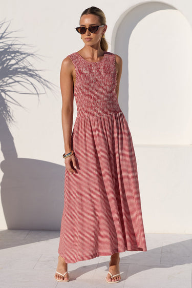 A woman modeling a sleeveless midi dress with a textured, smocked bodice and a fine vertical stripe pattern on the skirt, set against a bright white Mediterranean-style wall with an archway and a large terracotta pot.