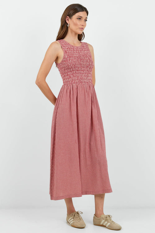 A woman modeling a sleeveless red mini gingham midi dress with a smocked bodice and side pockets, paired with beige sneakers, against a plain white studio background.