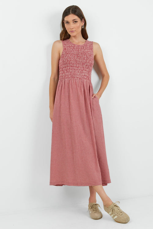 A woman modeling a sleeveless red mini gingham midi dress with a smocked bodice and side pockets, paired with beige sneakers, against a plain white studio background.
