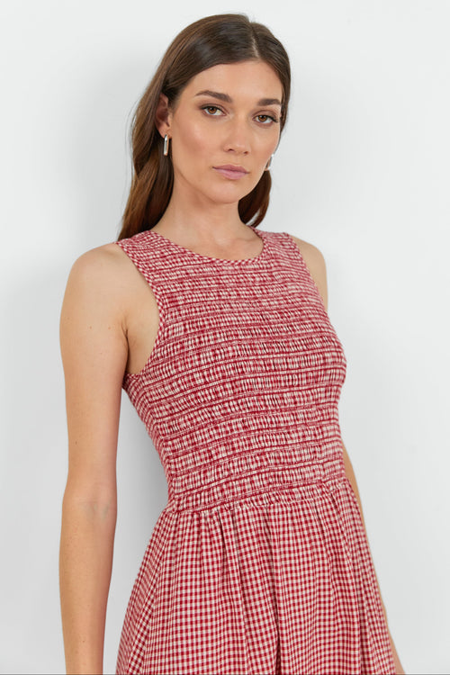 A woman wearing a sleeveless red and white gingham dress with a smocked bodice, standing against a plain light gray background.