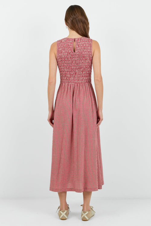 A woman modeling a sleeveless red mini gingham midi dress with a smocked bodice and side pockets, paired with beige sneakers, against a plain white studio background.