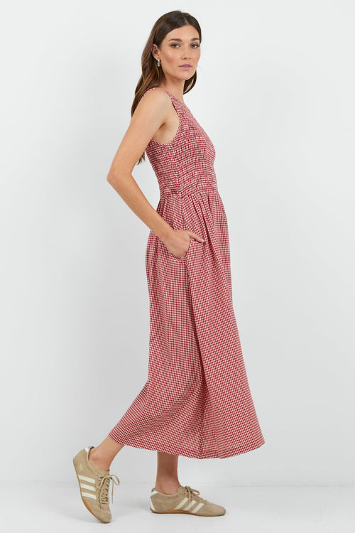 A woman modeling a sleeveless red mini gingham midi dress with a smocked bodice and side pockets, paired with beige sneakers, against a plain white studio background.