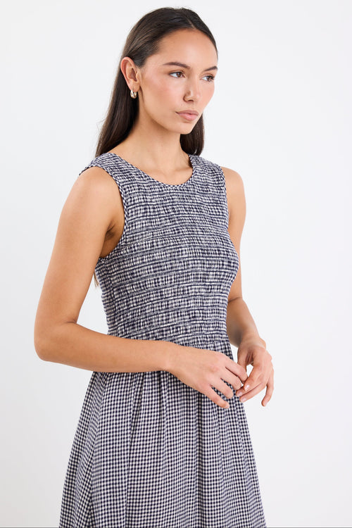 Woman wearing a navy mini gingham midi dress with a shirred bodice, sleeveless crew neckline, and relaxed gathered skirt.