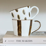Two stacked white ceramic mugs with black and gold speckled patterns, displayed on a stack of books on a white shelf.
