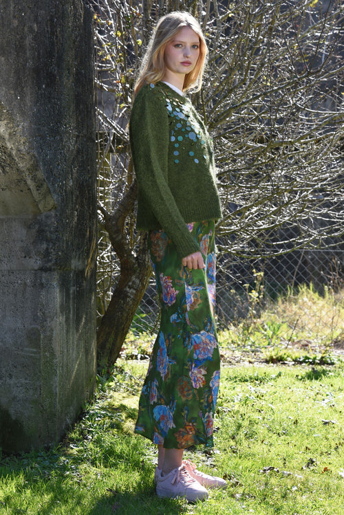 Woman wearing a green floral dress and sweater standing outdoors near a stone wall.