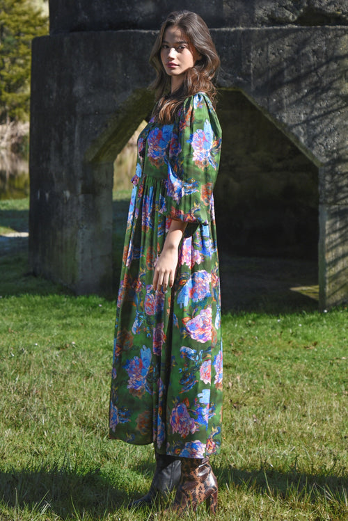 Woman wearing a long floral dress standing outdoors near a stone wall.