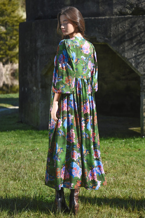 Woman wearing a colorful floral dress standing in a grassy area with a stone wall in the background.
