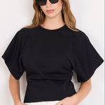 A woman modeling a black short-sleeve crewneck t-shirt paired with white pants and black sunglasses, standing against a plain white background.