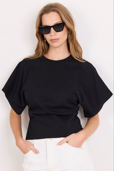 A woman modeling a black short-sleeve crewneck t-shirt paired with white pants and black sunglasses, standing against a plain white background.