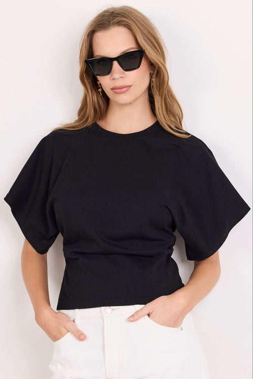 A woman modeling a black short-sleeve crewneck t-shirt paired with white pants and black sunglasses, standing against a plain white background.
