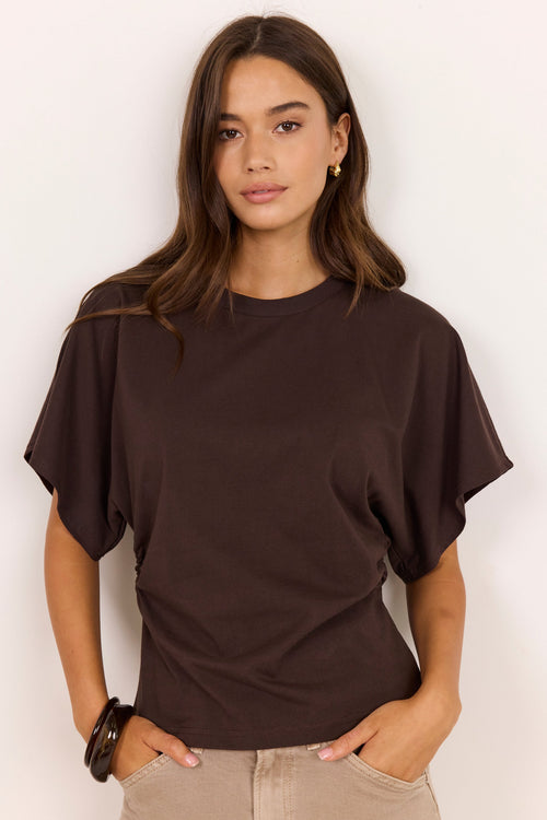 A woman wearing a dark brown short-sleeve t-shirt with draped, balloon sleeves and beige pants, standing against a plain off-white background.