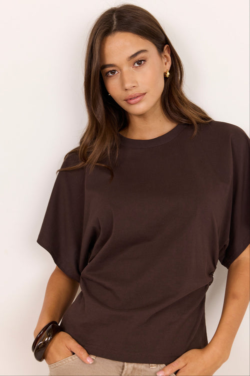 A woman wearing a dark brown short-sleeve t-shirt with a relaxed, draped fit, paired with beige pants, set against a plain off-white background.