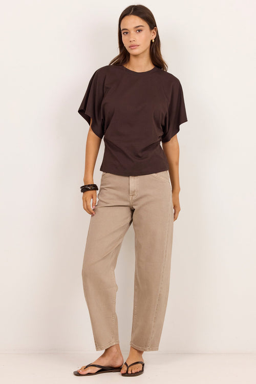 A woman modeling a dark brown short-sleeve t-shirt and beige straight-leg pants against a clean white studio background.