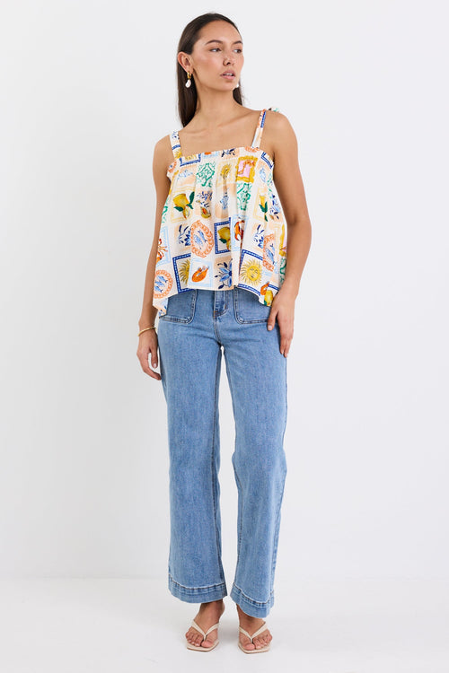 Woman wearing a colourful printed babydoll cami with tie straps, shirred neckline, and a loose, flowy fit paired with blue jeans.